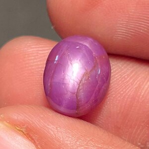 Puede incluir: Una gema rosa pulida, de forma ovalada, con efecto estrella. La gema se sostiene entre los dedos, mostrando su superficie lisa y patrones internos en forma de estrella. El color de la piedra varía de rosa claro a oscuro.