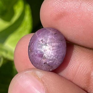 Op de afbeelding: Een kleine, ovaalvormige, paarsroze edelsteen. De edelsteen heeft een stervormig effect over het oppervlak. De achtergrond heeft groen gebladerte. De edelsteen wordt tussen vingers gehouden.