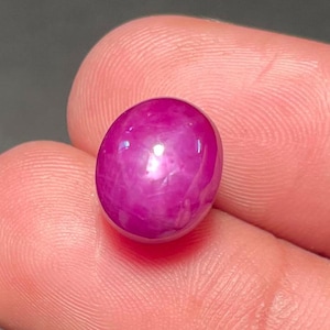 May include: A vibrant, oval-shaped ruby gemstone. The polished stone displays a deep, saturated pinkish-red hue with a smooth, reflective surface. The gemstone is held between fingers, showcasing its size and color.