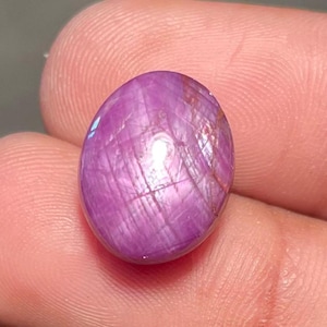 Puede incluir: Una gema pulida, de forma ovalada, de color rosa púrpura. La gema tiene una superficie lisa con líneas internas visibles y un brillo sutil. La piedra se sostiene entre dos dedos, mostrando su tamaño y color.