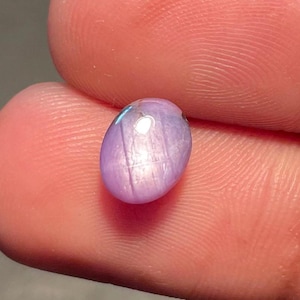 May include: A small, oval lavender gemstone. The polished surface reflects light, creating a star-like effect. The gem is held between two fingers, showcasing its size and colour. The gem is approximately 1 cm in length.