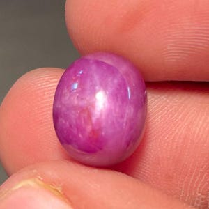 May include: A polished, oval-shaped ruby gemstone. The gem is a vibrant pinkish-red color with a star-like effect. The stone is held between two fingers, showcasing its smooth, reflective surface. This gemstone is ideal for jewelry making.