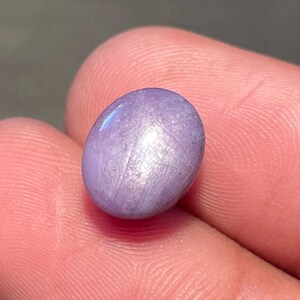 May include: A small, oval lavender gemstone. The polished surface reflects light, creating a subtle shimmer. The gem is held between two fingers, showcasing its size and colour. The stone's smooth texture and soft hue suggest it could be a cabochon.