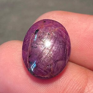 Puede incluir: Una gema de rubí pulida, de forma ovalada. La gema es de un rojo intenso con sutiles líneas internas y una superficie reflectante. El rubí se sostiene entre los dedos, mostrando su tamaño y color.