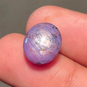 Può includere: Una gemma ovale lucidata con effetto stella. La gemma è di colore viola chiaro con un motivo a stella bianco al centro. La gemma è tenuta tra due dita.