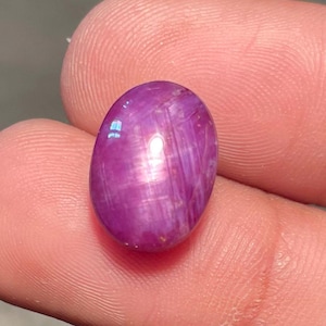 May include: A polished, oval gemstone in a deep magenta colour. The gem has a smooth surface with subtle internal lines, creating a star-like effect. The stone is held between fingers, showcasing its size and colour.