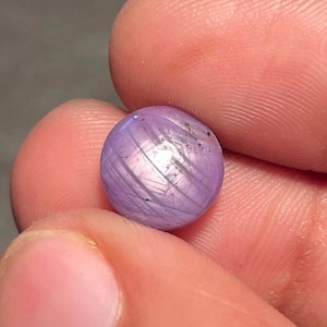 May include: A small, round, lavender-coloured gemstone with a star effect. The gem has a polished surface with visible lines and is held between fingers. The stone is a sphere, and the colour is a soft purple.