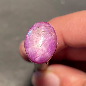 May include: A polished, oval-shaped gemstone with a vibrant pink hue. The gem has a star-like effect, with light reflecting across its surface. The stone is held between two fingers, showcasing its size and color.