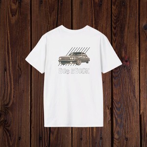 Puede incluir: Camiseta blanca con un gráfico de un coche clásico y el texto "80s 280CE". El coche es gris con líneas diagonales negras encima. La camiseta se muestra sobre un fondo de madera marrón oscuro.