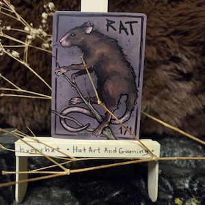 May include: A small, wooden easel displays a card featuring a brown rat illustration. The card has the word "RAT" at the top and the text "1/1" in the corner. The easel has the text "hxppy hxt • Hat Art And Gaming" on the front.