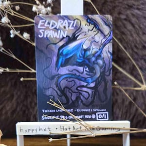 Könnte beinhalten: Eine rechteckige Spielkarte mit dem Titel Eldrazi Spawn und einer blauen und violetten Kreaturenillustration. Der Kartentext lautet Token Creature - Eldrazi Spawn, Sacrifice this creature: Add C und 0/1. Sie steht auf einer kleinen weißen Staffelei.