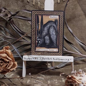 May include: A black and white illustration of a rabbit on a card with the word "Rabbit" at the top. The card is displayed on a small white easel. The text "Token Creature - Rabbit 1/1" is at the bottom of the card. The text "hxppy hxt • Hat Art And Gaming •" is on the easel.