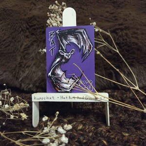 May include: A purple card featuring a bat illustration and the word "BAT" in white. The bat is rendered in black and white with red highlights. The card is displayed on a small white easel. The text "happy hxt • Hat Art and Games" is on the easel.
