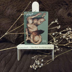 May include: A small, rectangular card featuring a detailed illustration of a wolf in shades of brown and black against a green background. The word "WOLF" is at the top. The card is displayed on a miniature white stand with the text "hxppy hxt • Hat Art And Gaming."