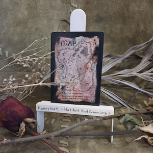 May include: A fantasy card with a hand-drawn map, displaying the word "MAP" at the top. The map is in brown and pink tones, with mountains and a river. The card is presented on a small white easel. Text at the bottom reads "hxpрy hxt • Hat Art And Gaming."