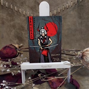 May include: A wooden art piece depicting a samurai warrior with a sword, set against a red sun and forest backdrop. The artwork includes the word "Samurai" and "Token Creature". The piece is displayed on a small white easel.