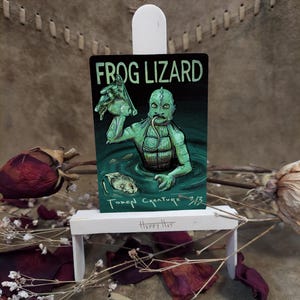 May include: A green and black illustration of a Frog Lizard Token Creature. The artwork depicts a creature emerging from water, with the text "FROG LIZARD" at the top. The card is displayed on a small white easel.
