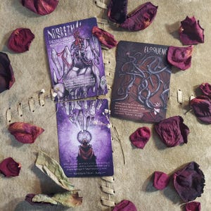 May include: Three fantasy-themed trading cards with illustrations and text, including "NASRETH THE SOUL HARROWER" and "ELOQUENC". The cards are on a textured surface, surrounded by dried rose petals. The cards have a purple and red color scheme.