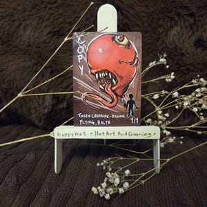 May include: A fantasy art card featuring a red balloon creature with sharp teeth and a long tongue. The card reads "COPY" and "TOKEN CREATURE - BALLOON FLYING, HASTE". A small silhouette of a person is also on the card. The card is displayed on a small white easel.