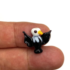 May include: A small glass figurine of a bald eagle with black wings, a white head, and a yellow beak. The eagle is perched on a finger.