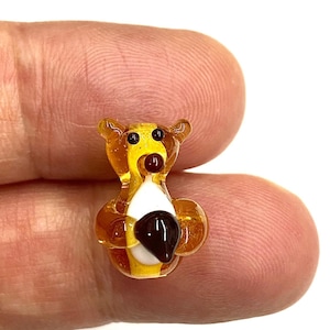 Puede incluir: Una pequeña figura de oso de cristal. El oso es principalmente de color ámbar con una cara amarilla y un pecho blanco. Tiene una nariz marrón oscuro y un cuerpo redondeado. La figura se sostiene entre dos dedos.