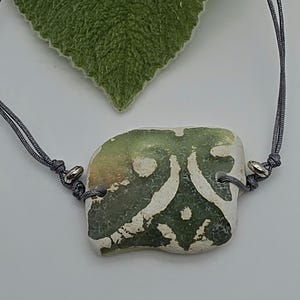 May include: A bracelet featuring a central, irregularly shaped piece of sea glass with a green and white patterned design. The glass is attached to a thin, dark gray cord with an adjustable sliding knot closure and small silver-toned metal beads.