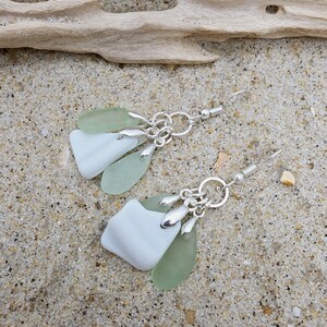 May include: A pair of dangle earrings featuring smooth, frosted white and pale seafoam green sea glass pieces suspended from silver-toned metal jump rings and fishhook wires. The design incorporates multiple layered glass shards.