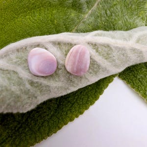 May include: Two smooth, polished, oval-shaped stones with a soft lavender and white banded pattern. The stones have a glossy finish and exhibit subtle, concentric color variations across their surfaces, displaying a light purple and white hue.