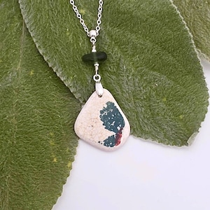 May include: A silver chain necklace featuring a pendant made from a smooth, triangular piece of off-white ceramic with a dark green and red speckled pattern. A small, dark green, rounded bead is positioned above the ceramic pendant.