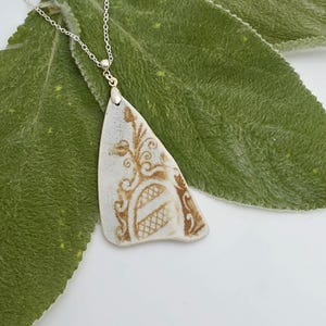 May include: A pendant necklace featuring a white ceramic shard with a brown decorative floral and lattice pattern. The shard is suspended from a thin silver-toned metal chain, which attaches to the top of the ceramic piece with a small metal bail.