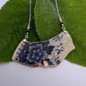 May include: A necklace featuring a curved, irregular ceramic shard pendant with a blue and white floral pattern and a crackled glaze finish. The pendant is suspended from a silver-toned metal chain accented with small white, black, and gray beads.
