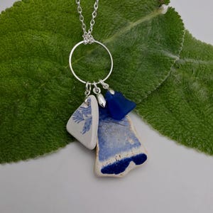 May include: A silver chain necklace featuring a circular pendant charm holder. Attached to the ring are three distinct charms: a white ceramic shard with blue floral patterns, a larger blue and white pottery fragment, and a small cobalt blue glass piece.
