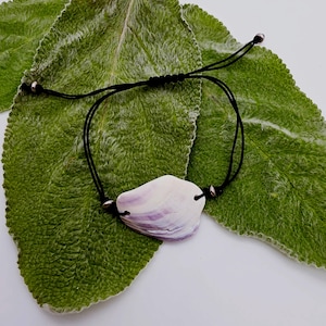 May include: A bracelet featuring a single, irregularly shaped seashell with white and purple hues, strung on a thin black cord. The cord includes an adjustable sliding knot mechanism and small metallic beads on either side of the shell.