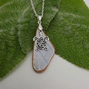 May include: A silver-toned metal turtle charm pendant attached to a triangular piece of smooth, light gray stone with a brown edge. The pendant hangs from a silver-toned metal chain, featuring an intricate filigree pattern on the turtle shell.