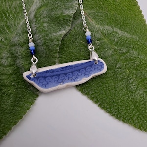 May include: A necklace featuring a rectangular piece of white and blue patterned ceramic pottery shard as a pendant. The pendant is suspended from a silver-toned metal chain, accented with small white, light blue, and dark blue glass beads.