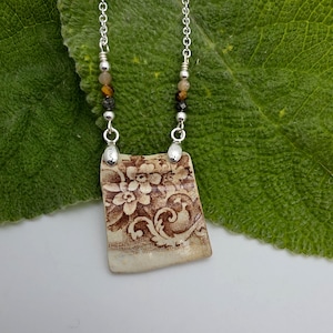 May include: A necklace featuring a rectangular ceramic pendant with a brown floral pattern on a cream background. The silver chain is accented with small, multi-colored beads including brown, black, and tan stones, connected by silver metal findings.