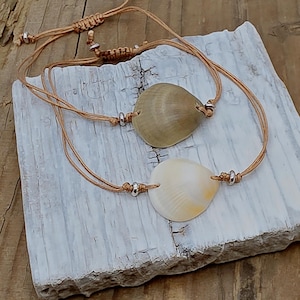 May include: Two seashell bracelets. One bracelet features a light brown shell, the other a white shell with yellow accents. Both shells are strung on tan cords with adjustable closures and silver-colored beads.