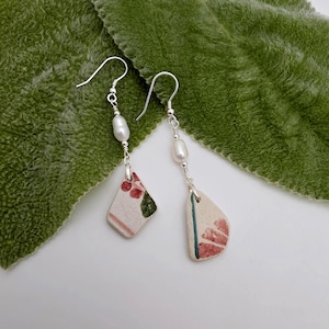 May include: A pair of drop earrings featuring silver-toned hooks, each with a single white freshwater pearl attached to a dangling piece of off-white ceramic pottery shard. The ceramic shards are decorated with small red floral patterns and green accents.
