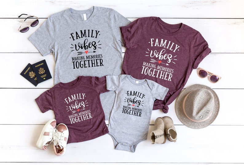 May include: A set of four shirts with the text "FAMILY vibes MAKING MEMORIES TOGETHER" in black and white. The shirts are in different sizes and colors, including gray and maroon. The shirts are laid out on a white wooden surface with other accessories, including sunglasses, a hat, and shoes.