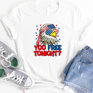 May include: White t-shirt with a cartoon bald eagle wearing sunglasses and a bandana with the American flag design. The eagle has a red beak and yellow eyes. The text "YOU FREE TONIGHT?" is printed below the eagle.