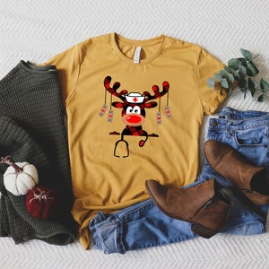 May include: Mustard yellow t-shirt featuring a cartoon reindeer wearing a nurse's cap and stethoscope. The reindeer has a red nose and antlers with a buffalo plaid pattern. The shirt is paired with blue jeans, brown boots, and a green sweater.
