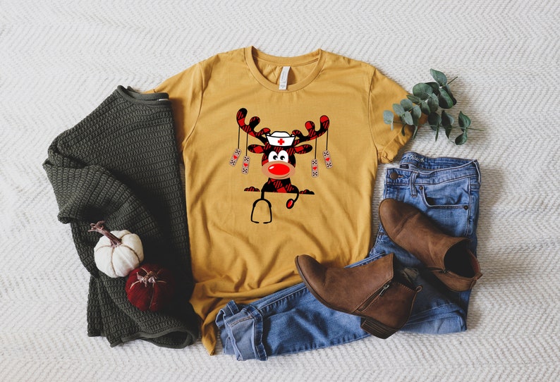 May include: Mustard yellow t-shirt featuring a cartoon reindeer wearing a nurse's cap and stethoscope. The reindeer has a red nose and antlers with a buffalo plaid pattern. The shirt is paired with blue jeans, brown boots, and a green sweater.
