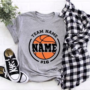 May include: Gray t-shirt with a black and orange basketball graphic. The basketball has the text "TEAM NAME" above it and "NAME" inside the basketball. The number "#16" is below the basketball.