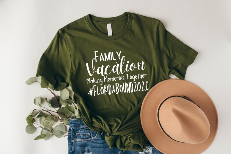 May include: A green t-shirt with the text "FAMILY vacation Making Memories Together #FLORIDABOUND2021" in white lettering. The shirt is paired with blue jeans and a brown hat.
