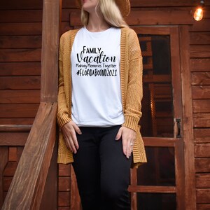May include: A white t-shirt with the text "FAMILY Vacation Making Memories Together #FLORIDABOUND2021" printed in black. The shirt is being worn by a person standing in front of a wooden door.