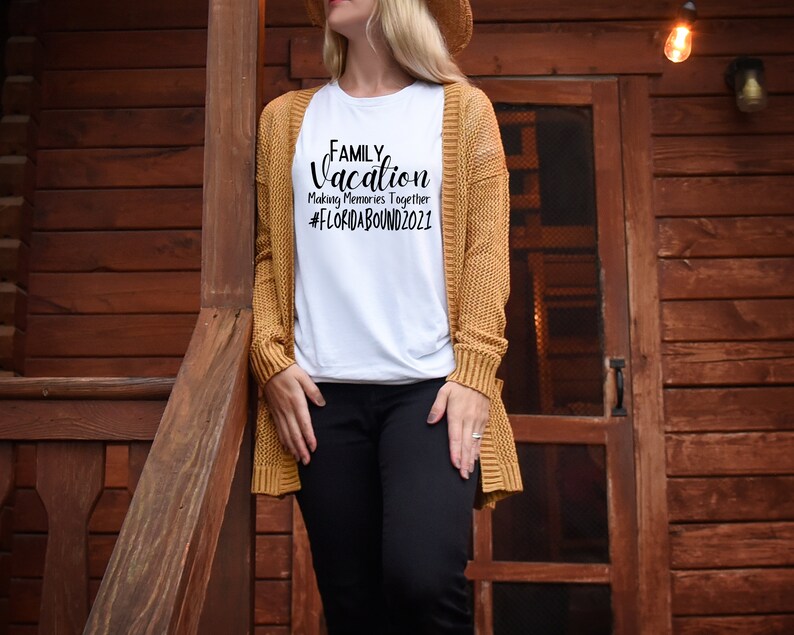 May include: A white t-shirt with the text "FAMILY Vacation Making Memories Together #FLORIDABOUND2021" printed in black. The shirt is being worn by a person standing in front of a wooden door.