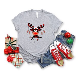 May include: A heather gray t-shirt featuring a Christmas reindeer design. The reindeer has a nurse's cap, a red nose, and a plaid pattern. A stethoscope is also part of the design. The shirt is paired with blue jeans and red sneakers.