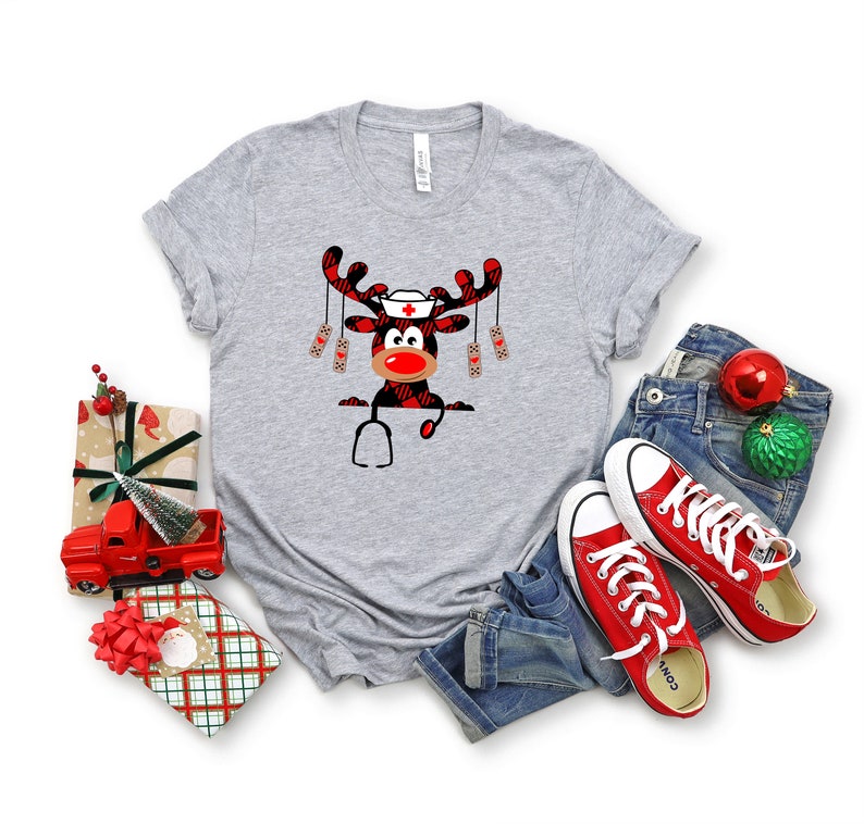 May include: A heather gray t-shirt featuring a Christmas reindeer design. The reindeer has a nurse's cap, a red nose, and a plaid pattern. A stethoscope is also part of the design. The shirt is paired with blue jeans and red sneakers.