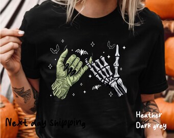 May include: Dark grey t-shirt featuring a graphic of two hands, one green and one skeletal, interlocked in a pinky promise. The green hand has long black fingernails. Bats and stars surround the hands. The text "Heather Dark grey" is visible.