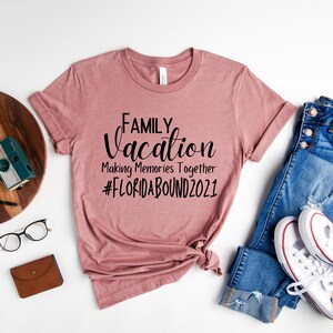 May include: A dusty rose t-shirt with the text "FAMILY Vacation Making Memories Together #FLORIDABOUND2021" printed in black. The shirt is laid flat on a white surface with a pair of blue jeans, white trainers, a wooden chopping board, a camera, a watch, and glasses.